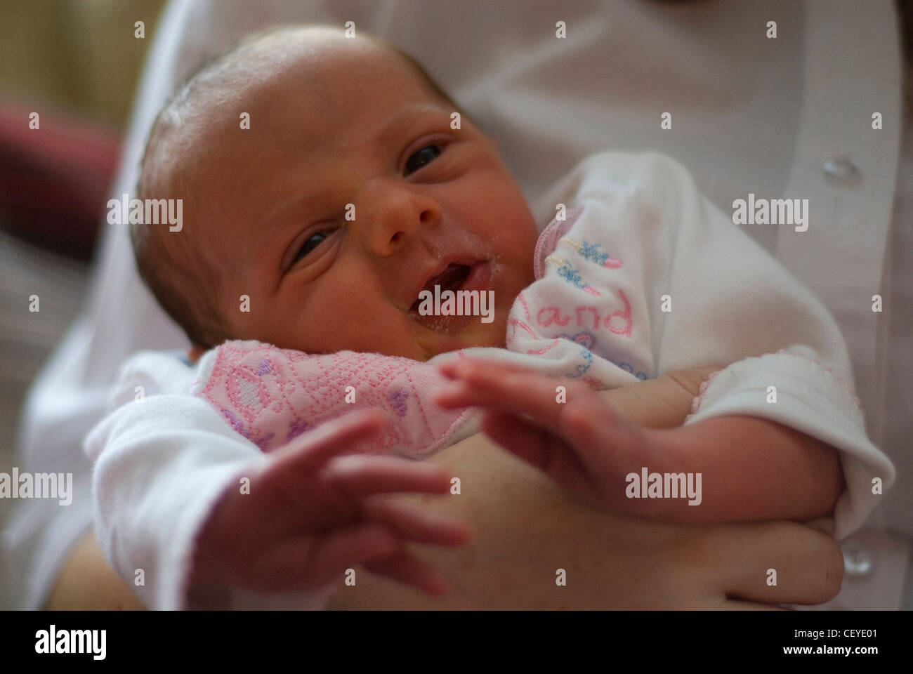 A newborn female baby dressed in a white patterned babygrow, his mouth open and looking at the camera, being held by an adult Stock Photo