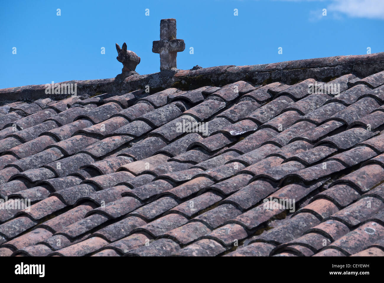 An old and weathered red ceramic tile roof detail of the Catholic church with stone cross and rabbit on the crest of the roof. Stock Photo