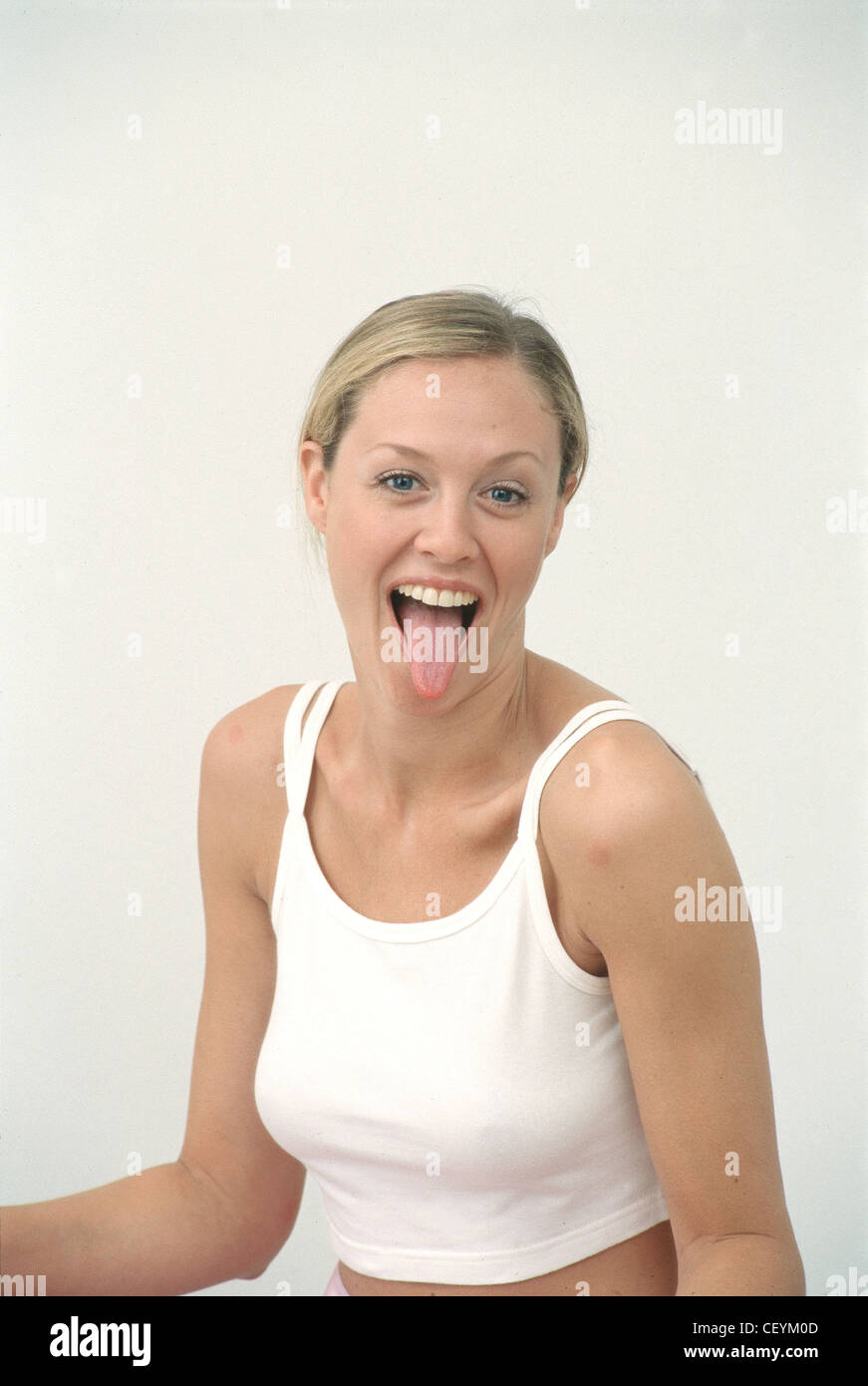 Female with long blonde hair off face wearing cropped white vest top and subtle make up looking to camera sticking tongue out Stock Photo