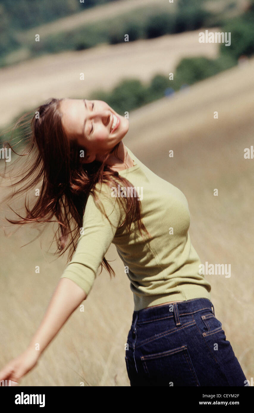Semi profile of female long brunette hair wearing subtle make up green top and blue jeans standing in field arms outstretched Stock Photo