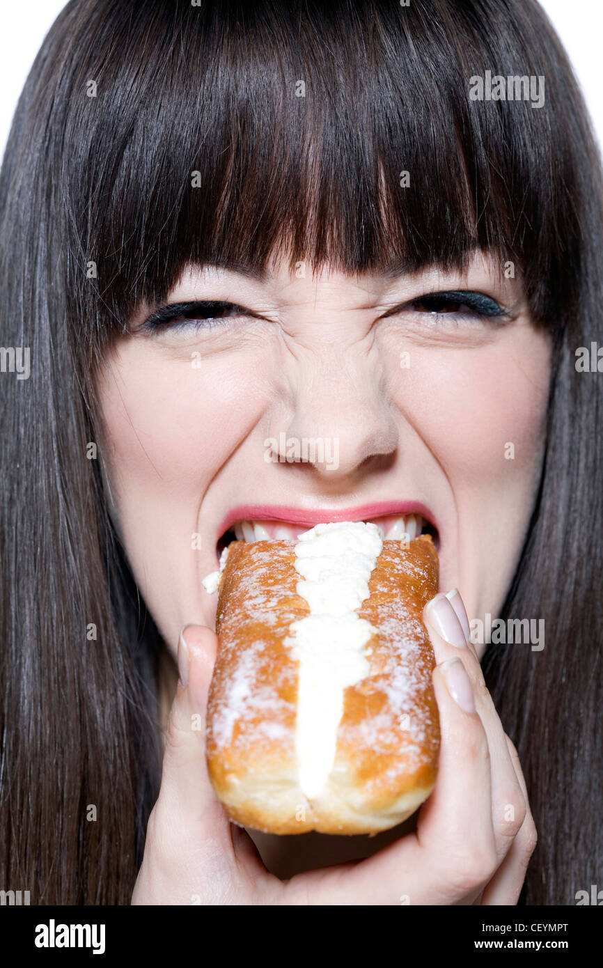 Female long fringed brunette hair, wearing green metallic eyeshadow and pink lipgloss, eating eclair, smiling, looking at Stock Photo