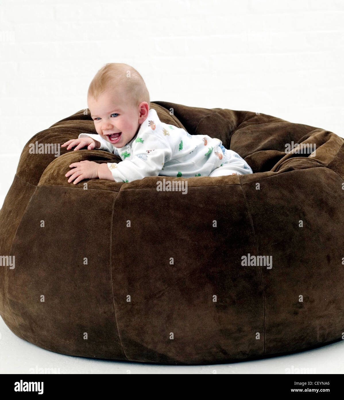 Male baby wearing white patterned babygro, lying on front on large brown suede beanbag looking to distance smiling, against Stock Photo