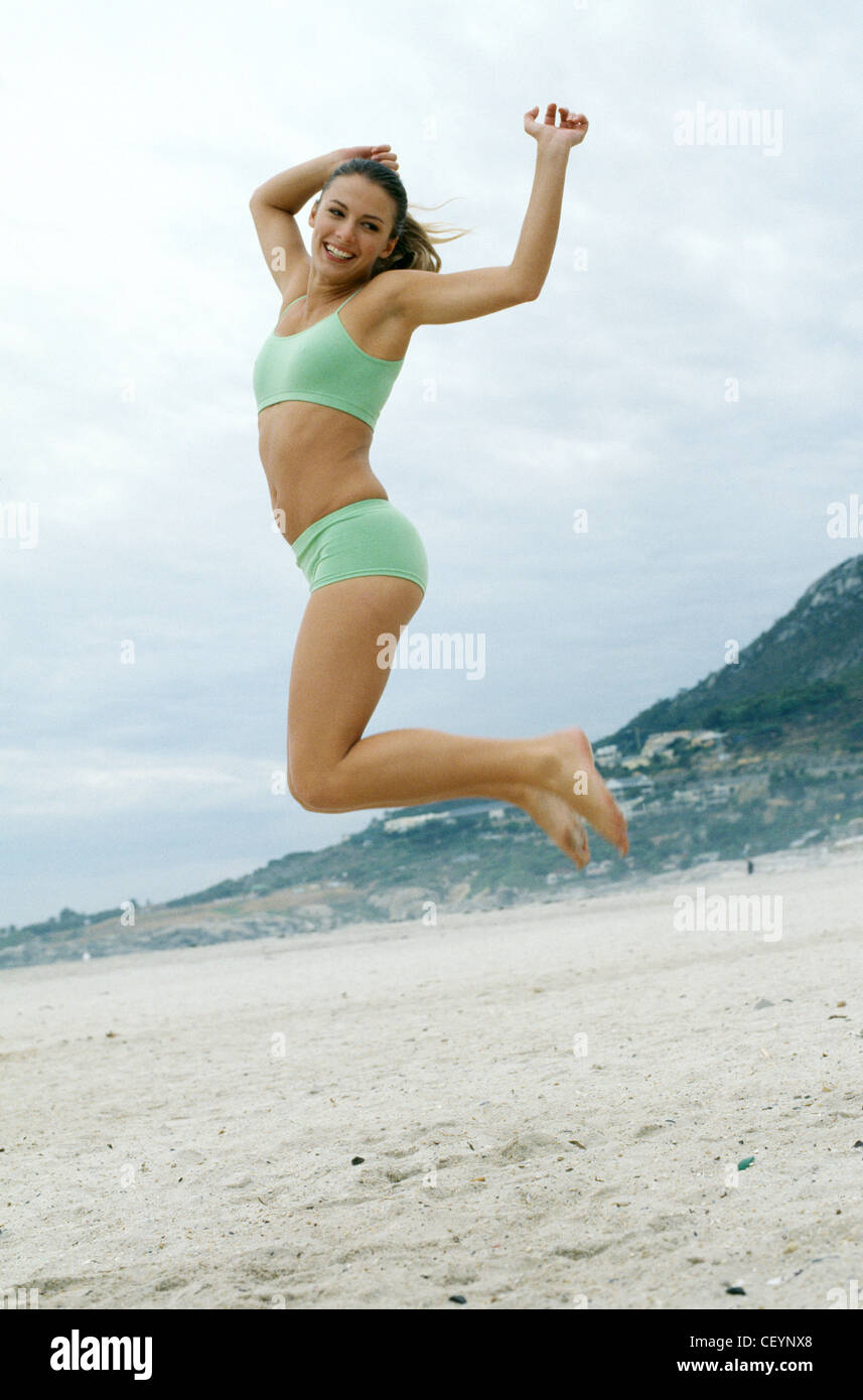 Smiling brunette female jumping up on secluded sandy beach, both arms up, both knees bent backwards, facing sideways, smiling Stock Photo
