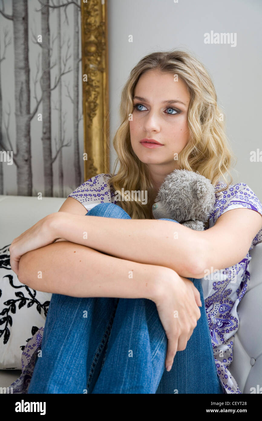 Female Long Blonde Wavy Hair Wearing Purple And White Printed Top And Blue Jeans Hugging Knees