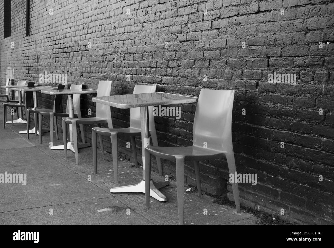 Black and white image of brushed aluminum tables and chairs on a sidewalk against a black brick wall Stock Photo