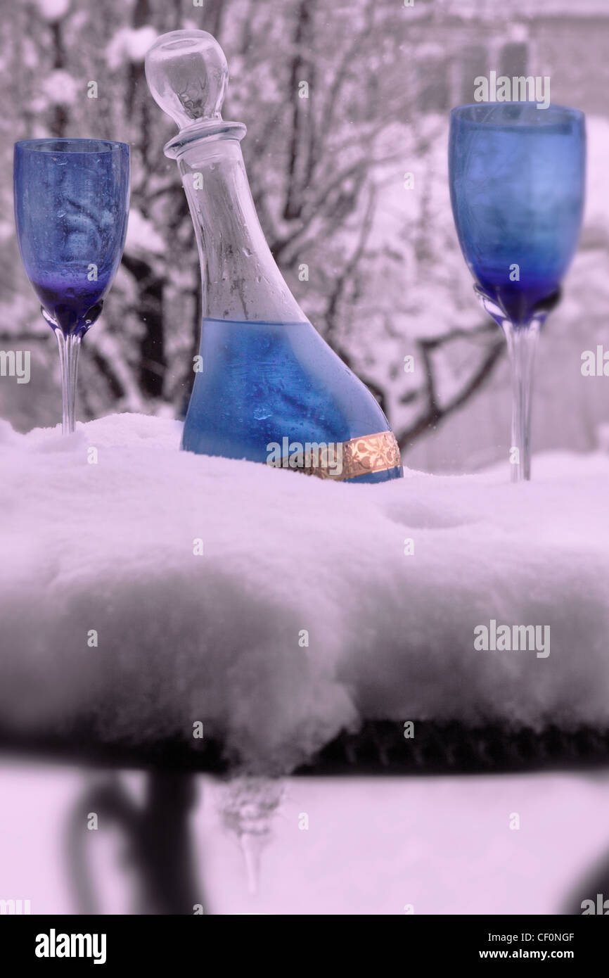 Outside it's snowing. On the snowy table, a decanter is filled with a cold bluish drink. Two blue glasses are part of the scene. Stock Photo