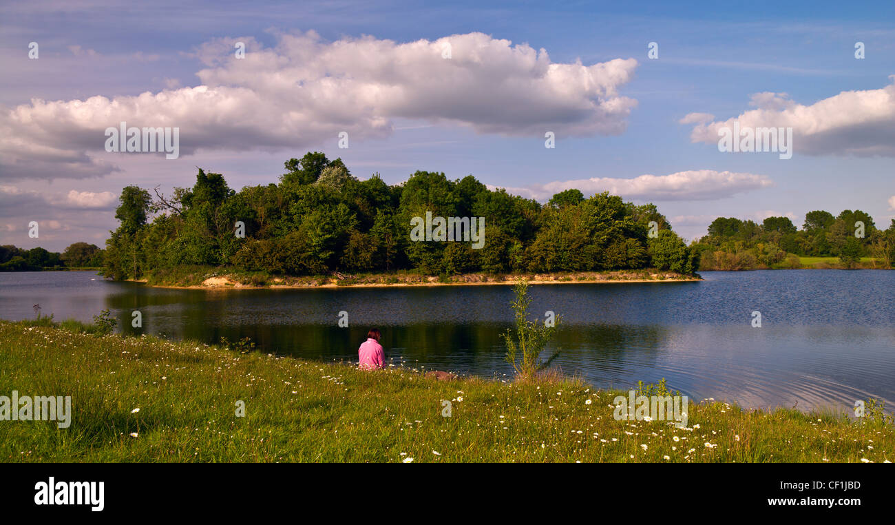 A woman sitting on a bank by the side of Lake 29 of the Cotswold Water Park. The Cotswold Water Park is formed from over 150 lak Stock Photo