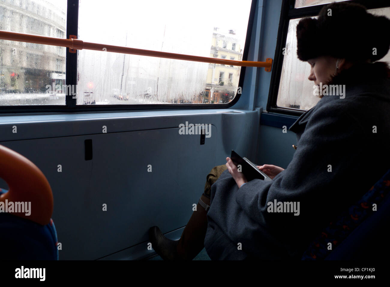 A fashionable young woman wearing a fur hat reading a Kindle electronic book sitting at the top front window of a double-decker bus  in London UK Stock Photo