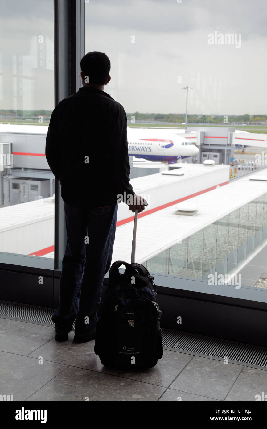 Silhouette of a passenger standing with his travel bag looking out of a window in the departure lounge of Heathrow Airport. Stock Photo
