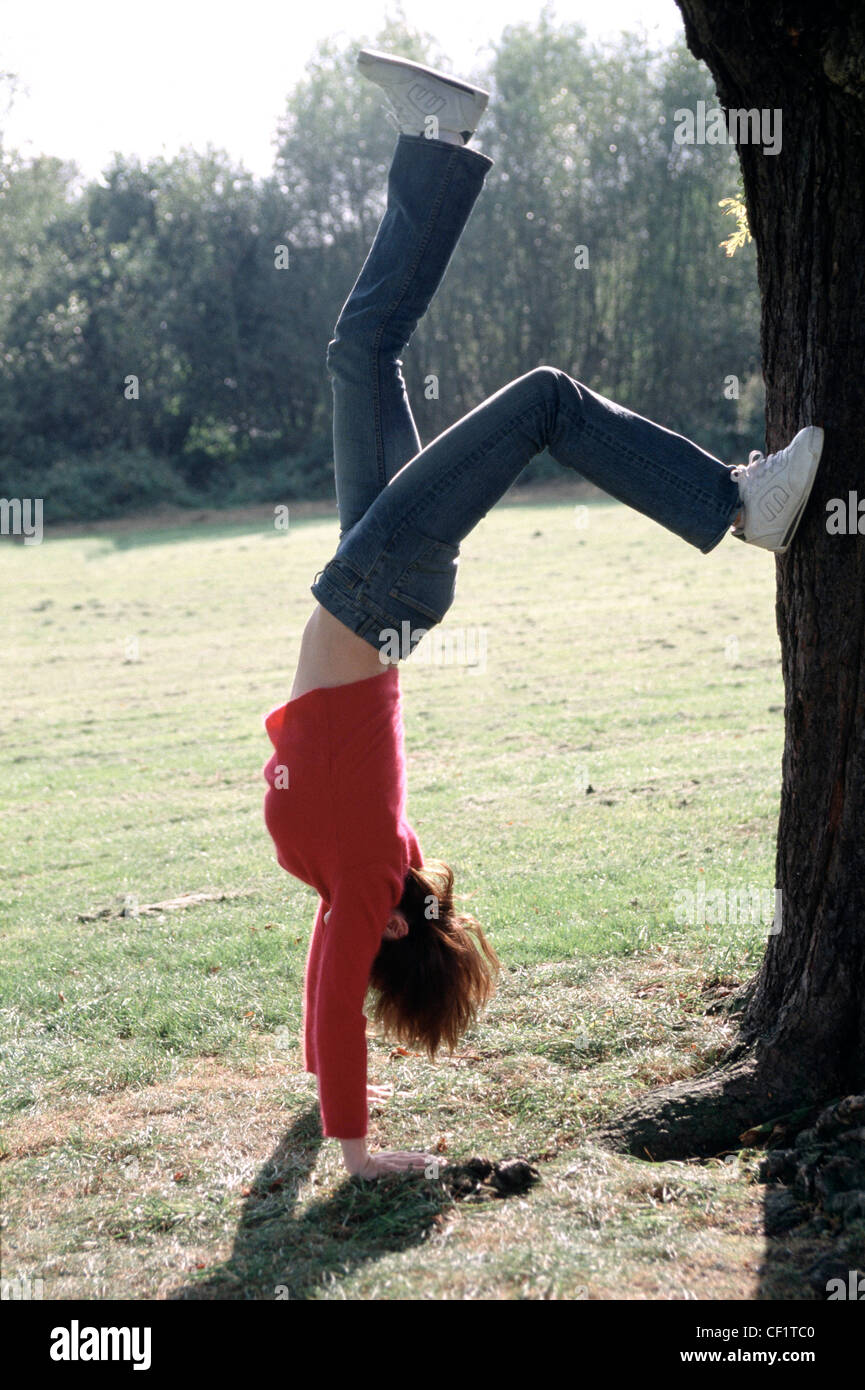 A Female short auburn hair wearing bright pink fluffy jumper blue jeans and trainers, doing a handstand in park, one foot on Stock Photo
