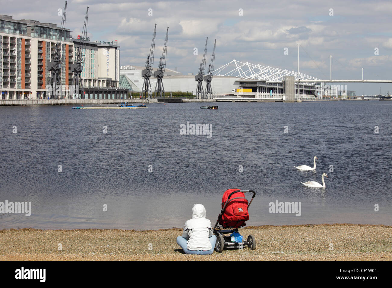 A mother and child in a push-chair sitting on a bank of the River Thames looking towards the Royal Victoria Docks Stock Photo