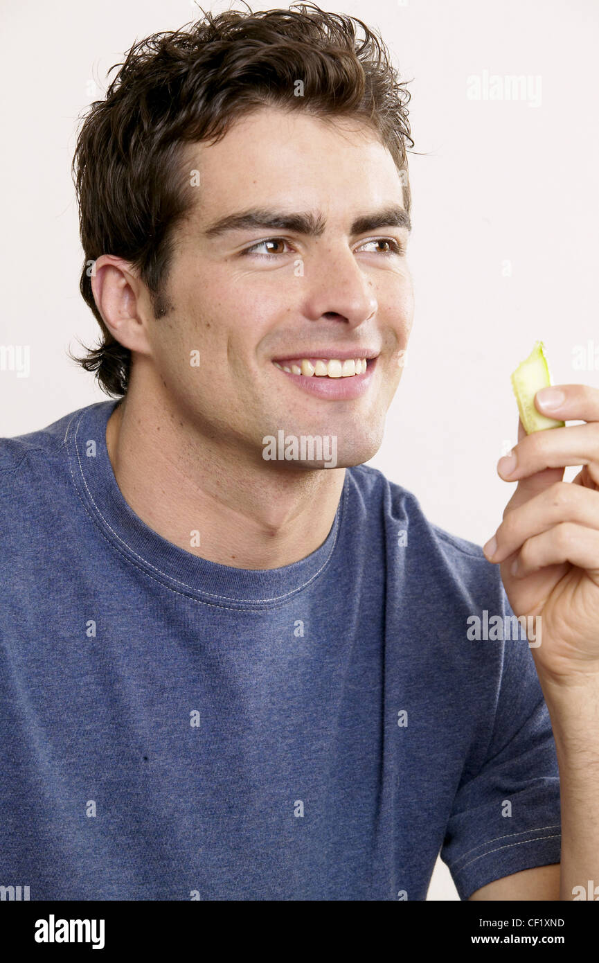 Male with dark brown hair wearing blue t shirt, holding piece of cucumber looking to side smiling, white wall in background Stock Photo