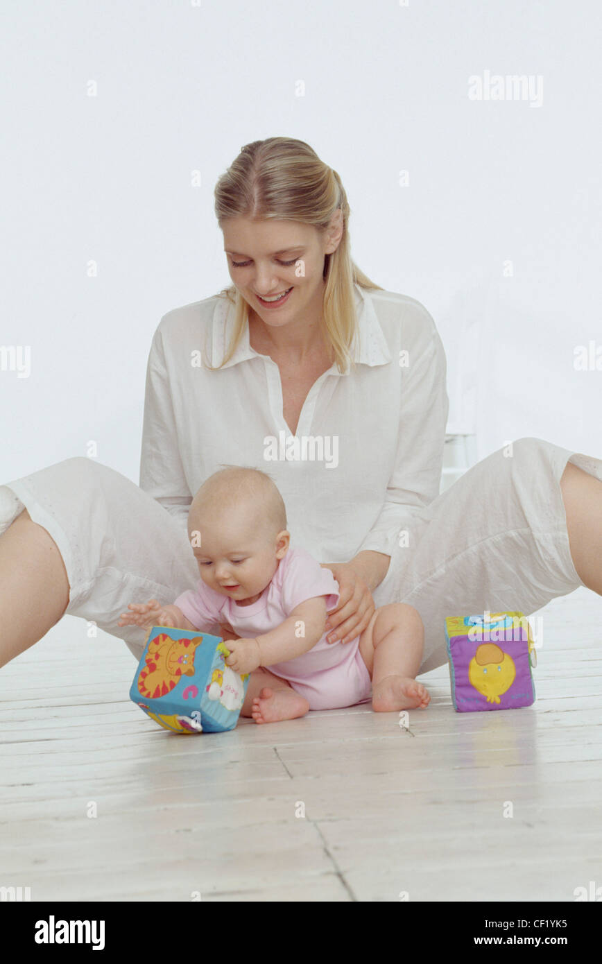 Female blonde hair off face wearing white blouse and white cropped trousers sitting on flowith legs wide open, looking down Stock Photo