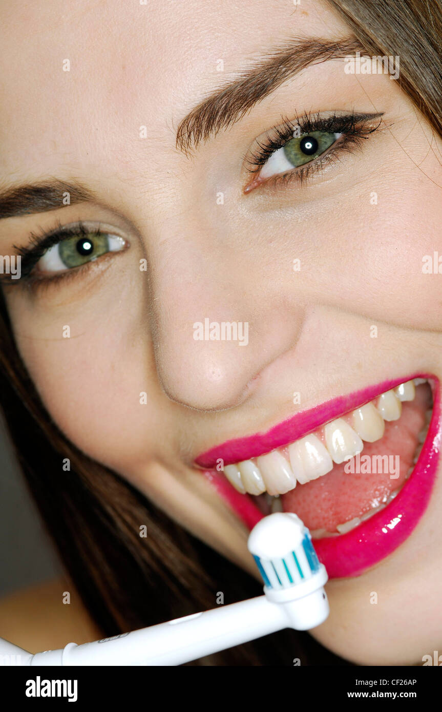 Close up of female brunette hair wearing black eyeliner, mascara and bright pink lipgloss holding electric toothbrush to mouth Stock Photo