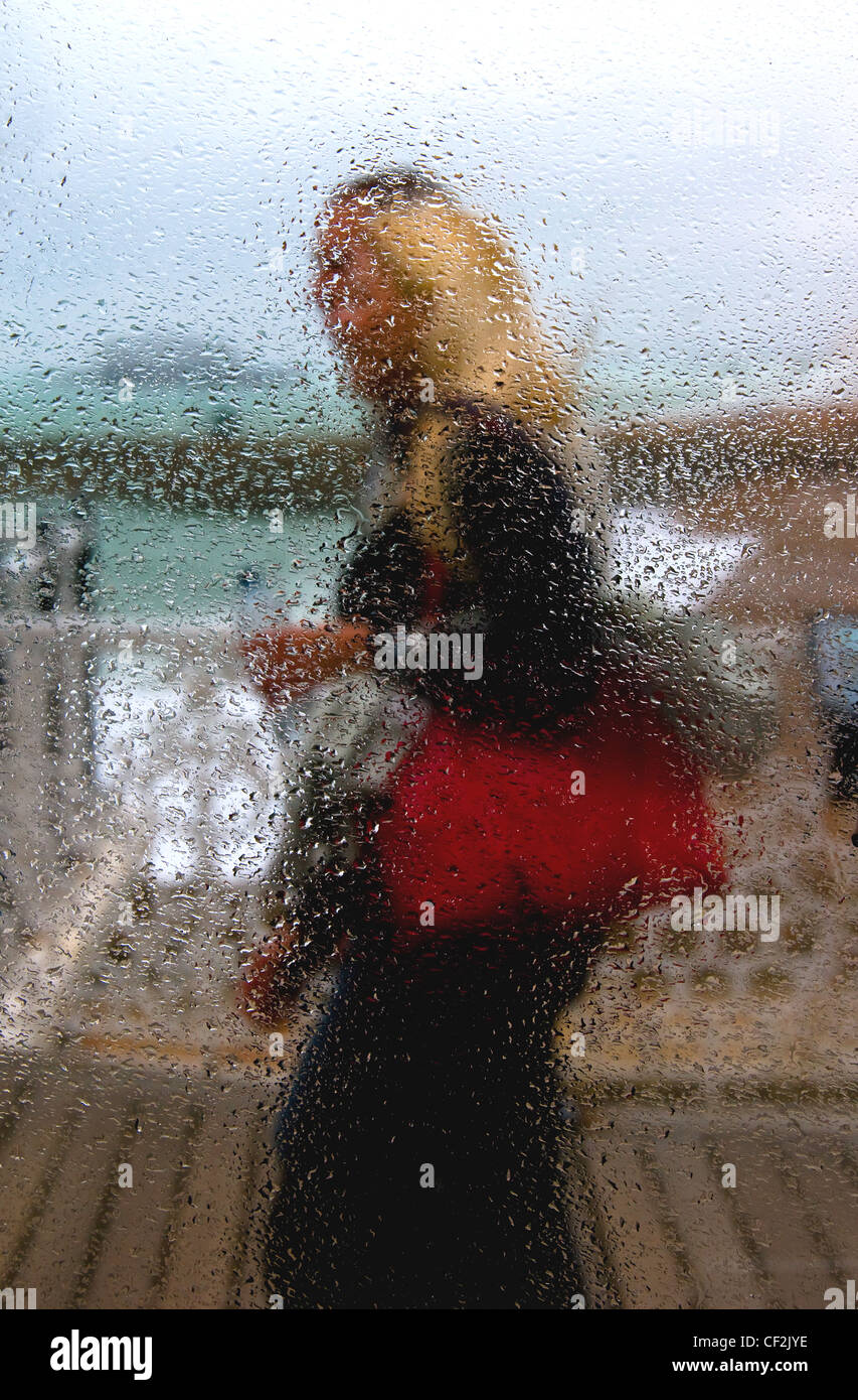 A couple seen through water droplets on a glass window on Brighton Pier on a rainy day. Stock Photo