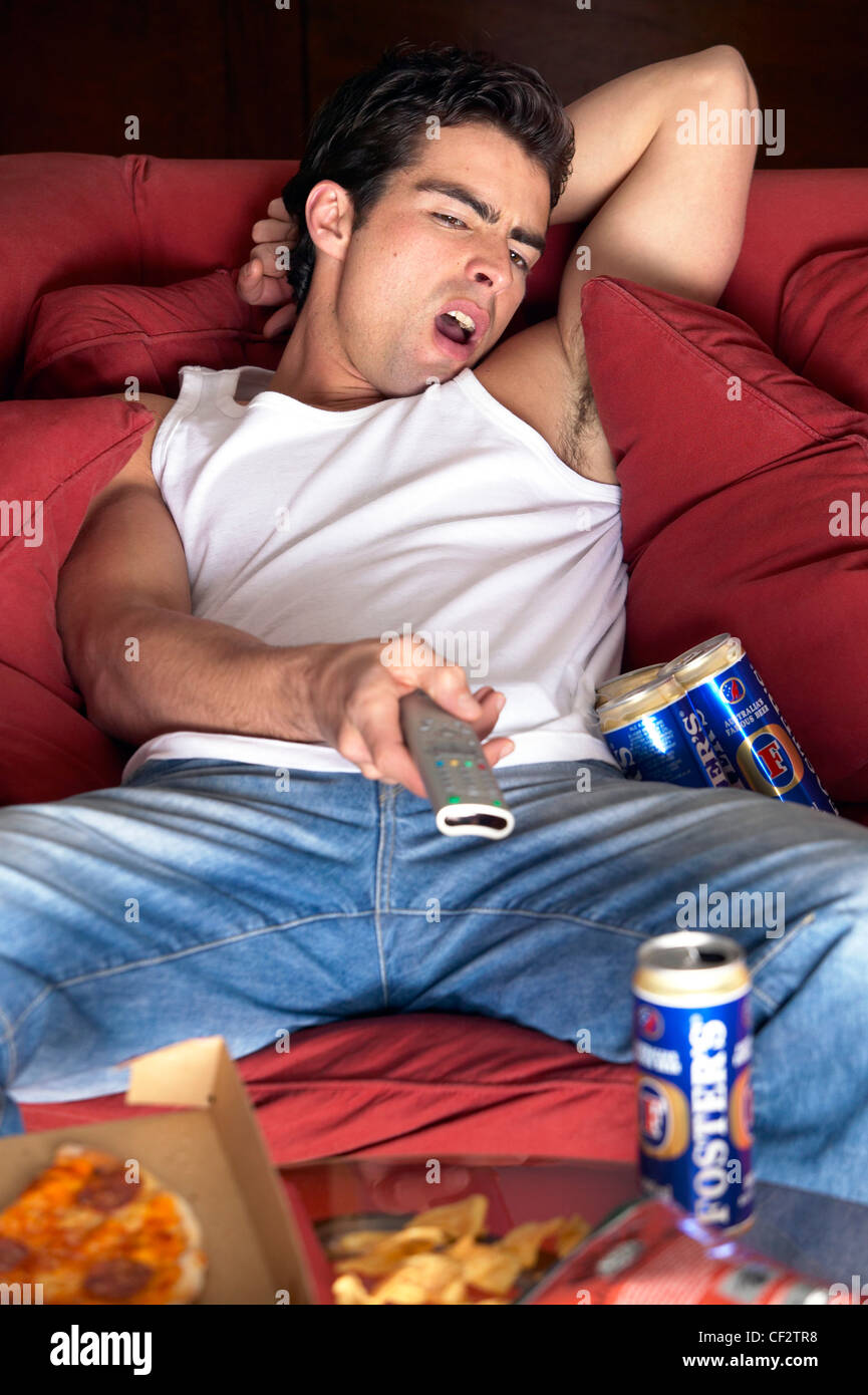 Male dark brown hair wearing white vest top and blue jeans, sitting slouched on red sofa watching tv one arm raised behind Stock Photo