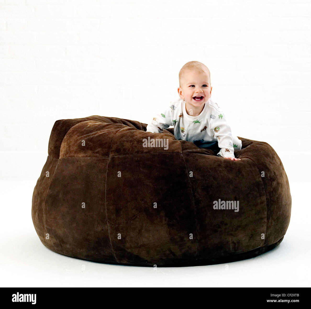 Male baby wearing white patterned babygro, crawling on large brown suede beanbag looking to distance smiling, against white Stock Photo