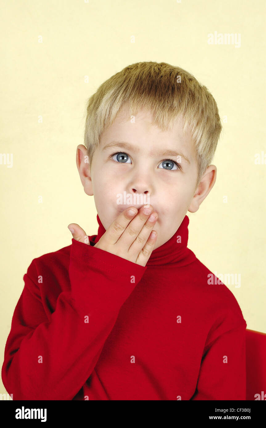 Blonde three year old boy wearing red turtle neck jumper hand covering his mouth Baba and Kleuter Stock Photo