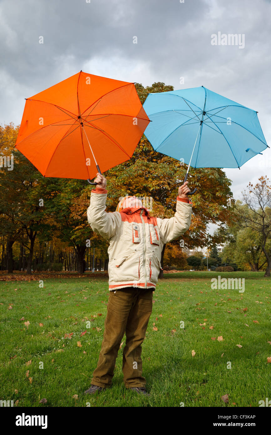 Boy in autumn park. Holds over head two colour umbrellas under cloudy sky. Stock Photo