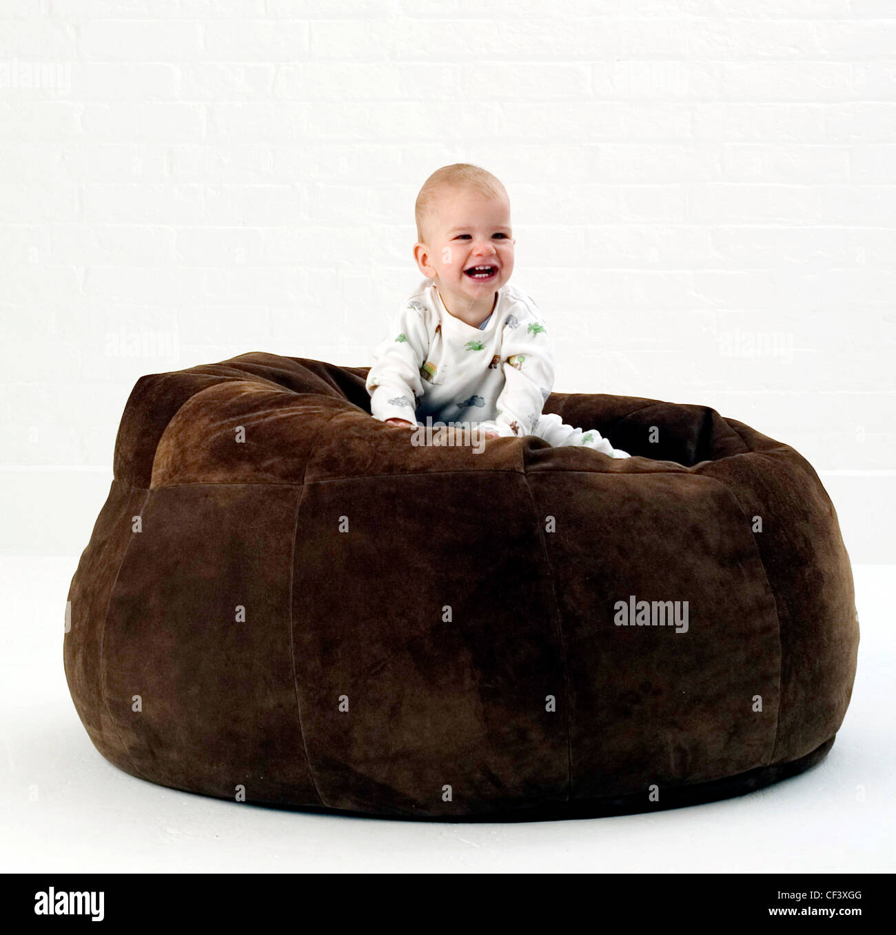 Male baby wearing white patterned babygro, sitting on large brown suede beanbag looking to distance smiling, against white Stock Photo