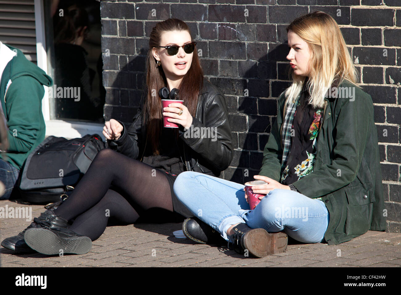 Portrait of two young women talking, drinking and sitting the pavement, London, England, UK. Stock Photo