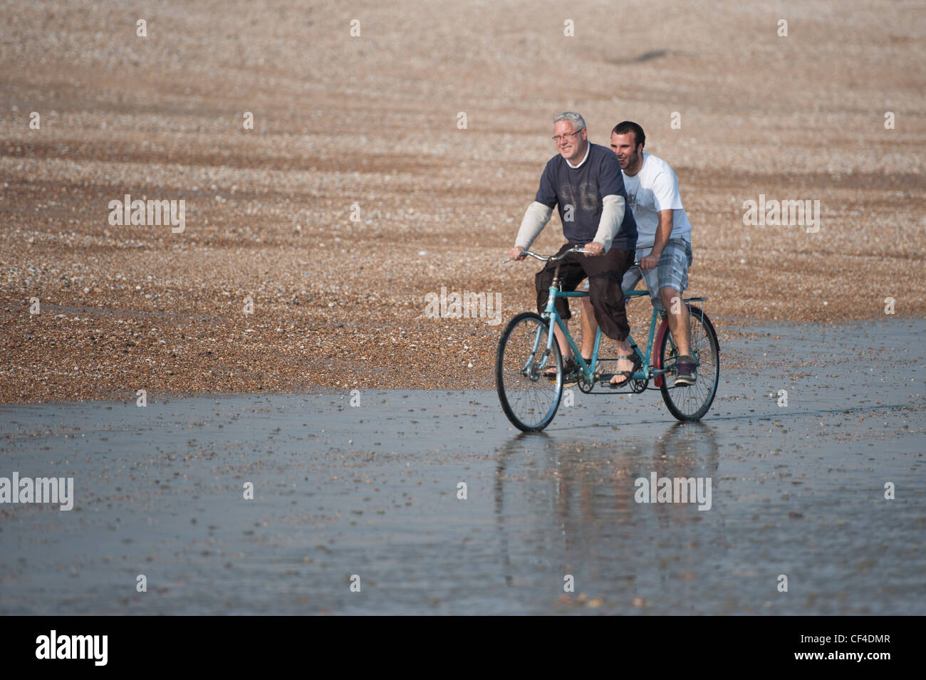 Two men riding on tandem along waters edge on beach Stock Photo