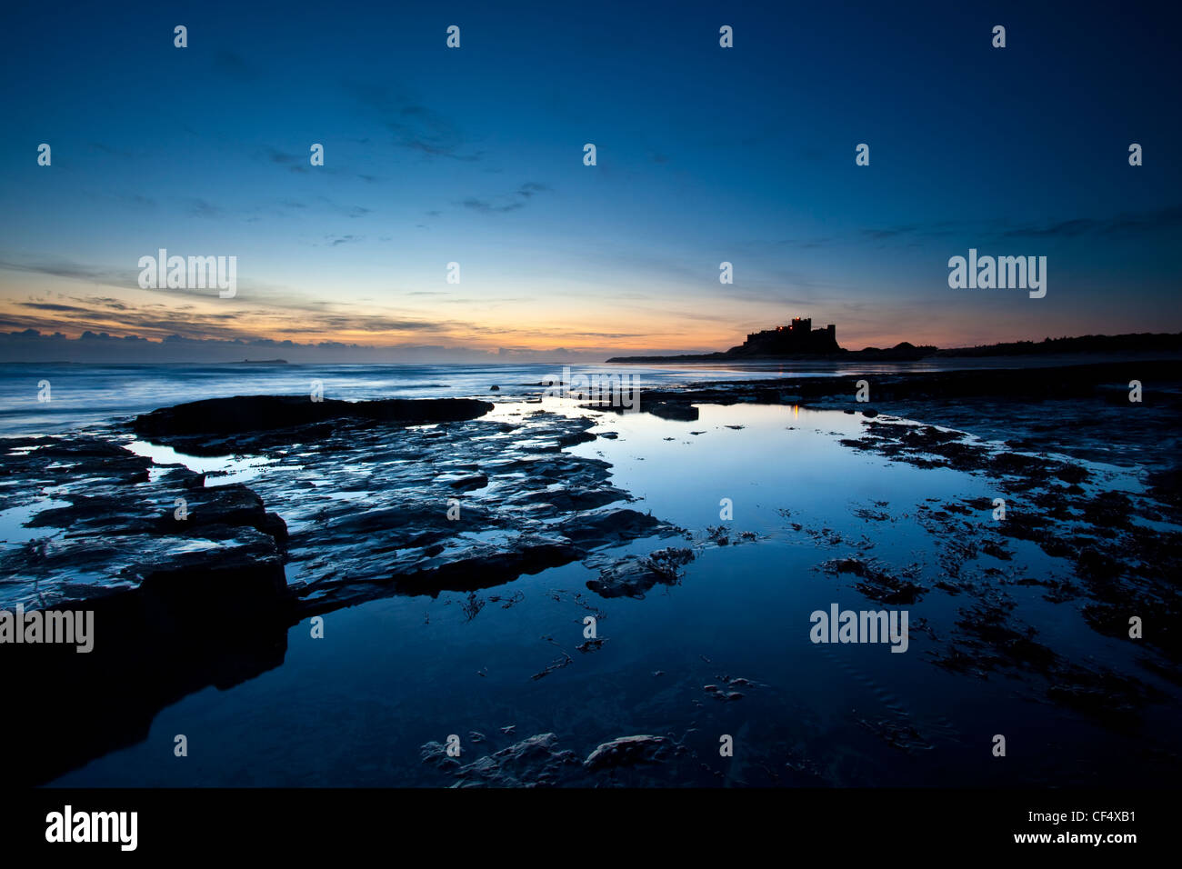 View across the rocky shore towards Bamburgh Castle silhouetted by early dawn light. Stock Photo