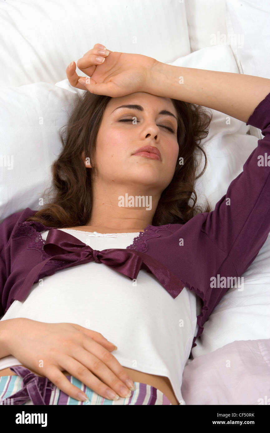 Winter Lifestyle Female shoulder length brunette hair laying on her back in bed, head on a white pillow One arm over stomach Stock Photo