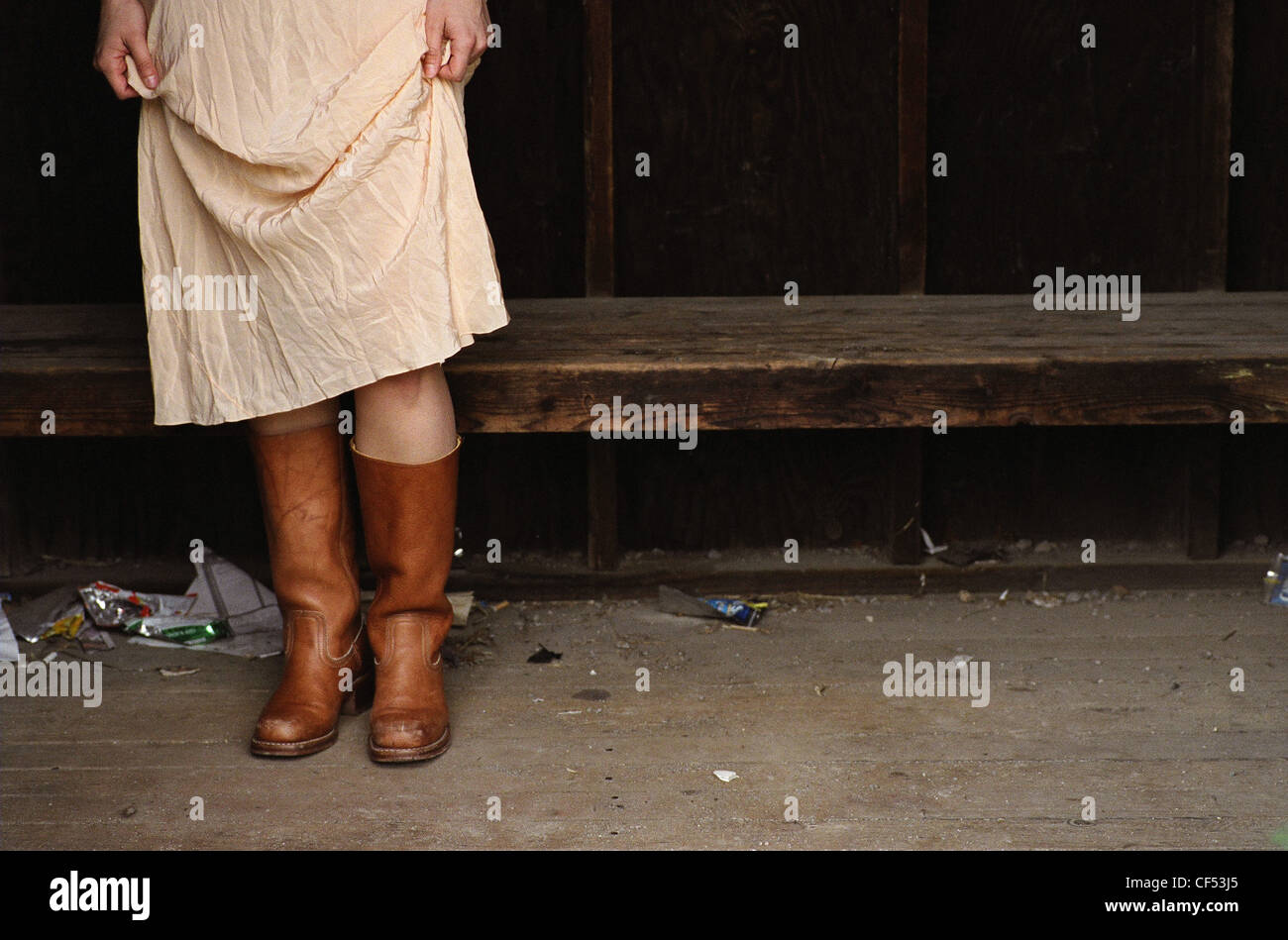 Female wearing crumpled peach dress tan leather boots Standing in a shelter by a wooden bench rubbish on the floor Slightly Stock Photo