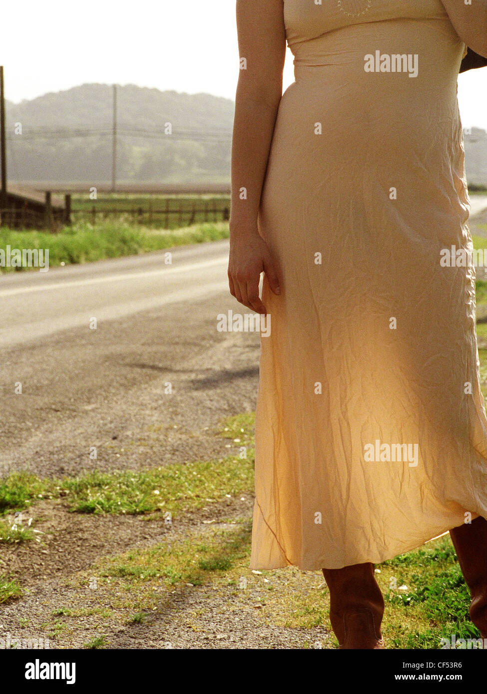 Female wearing crumpled peach dress tan leather boots Standing by the side of a road Rural scene in background Light showing Stock Photo