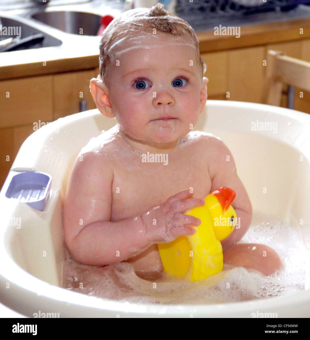 Female child sitting in baby bath covered in soap holding yellow rubber