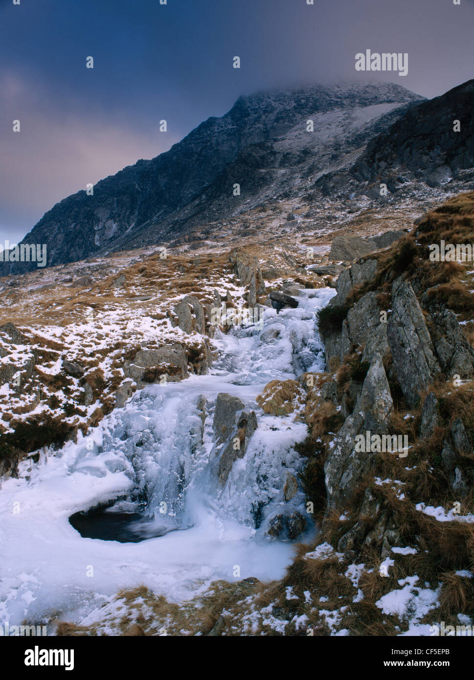 Small waterfall and partly frozen pool on a stream flowing down from Llyn Bochlwyd to Llyn Ogwen. The dark mass of Tryfan is bey Stock Photo