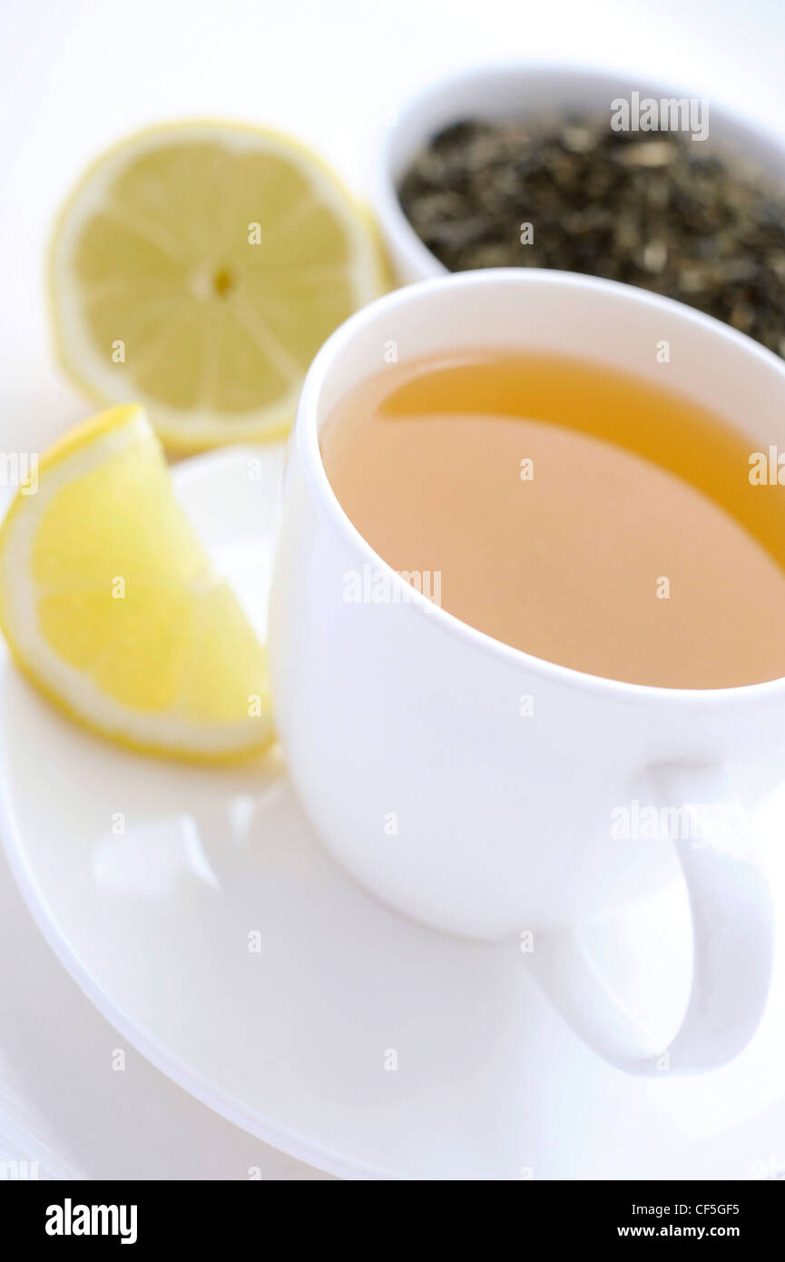 A close up of a white cup and saucer filled a yellow fruit tea, a slice of lemon on one side, the rest of the lemon and a Stock Photo
