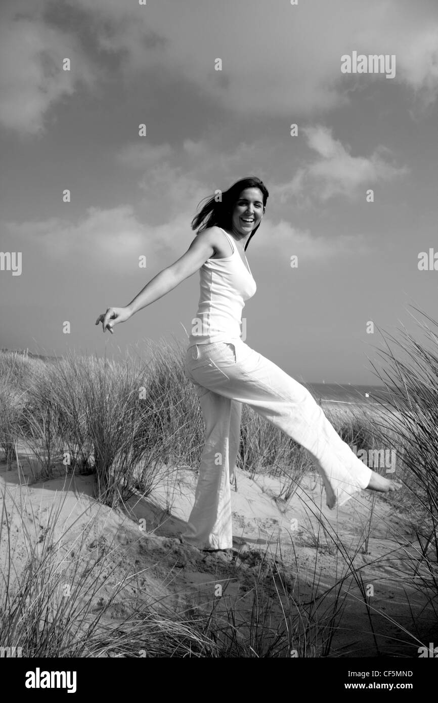Female shoulder length brunette hair wearing a white vest and white trousers, standing on the beach, one leg raised, arms Stock Photo