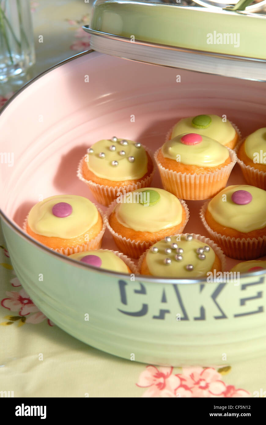 An enamel cake tin filled with various fairycakes, topped with cream coloured icing and various sweets Stock Photo