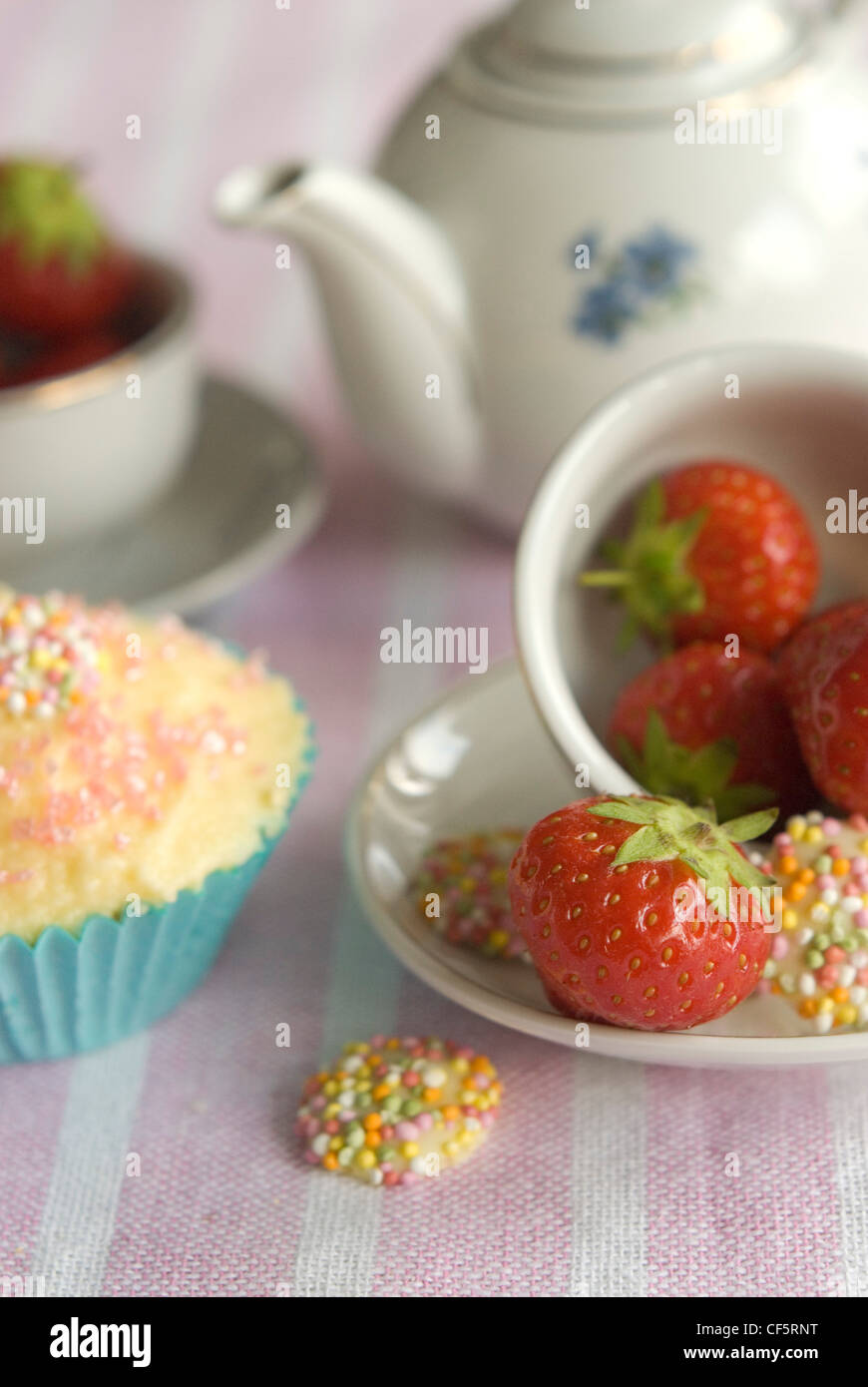 A close up of a strawberry white chocolate sweets in a toy china tea cup, fairy cakes butter icing and pink spinkles on a pink Stock Photo