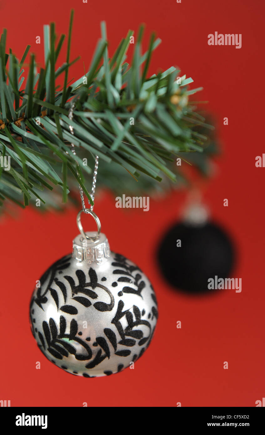 A metallic silver and black patterned bauble hanging off the branch of a christmas tree, a black bauble behind it, a red Stock Photo