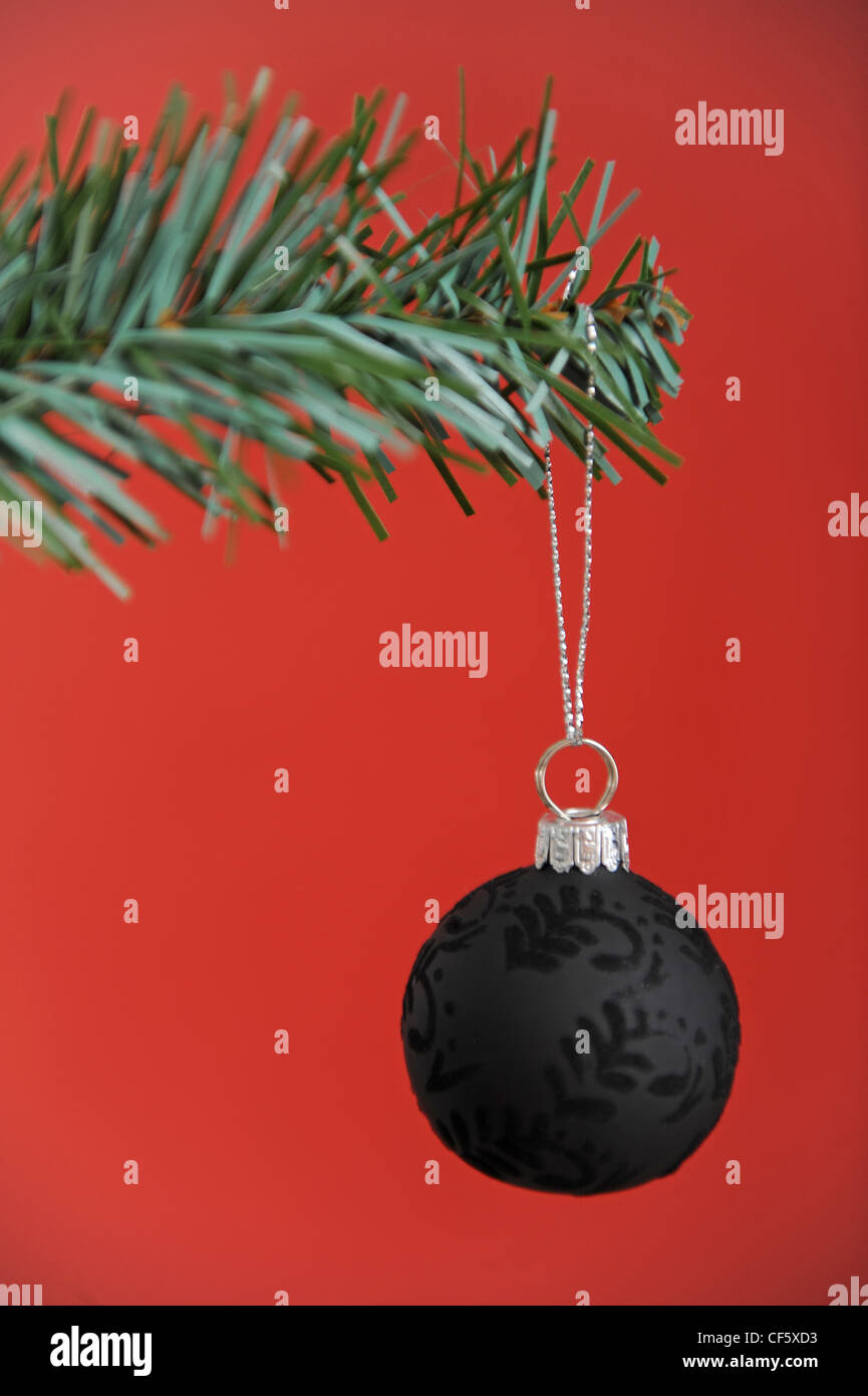 A black patterned bauble hanging off the branch of a christmas tree, with a red background Stock Photo