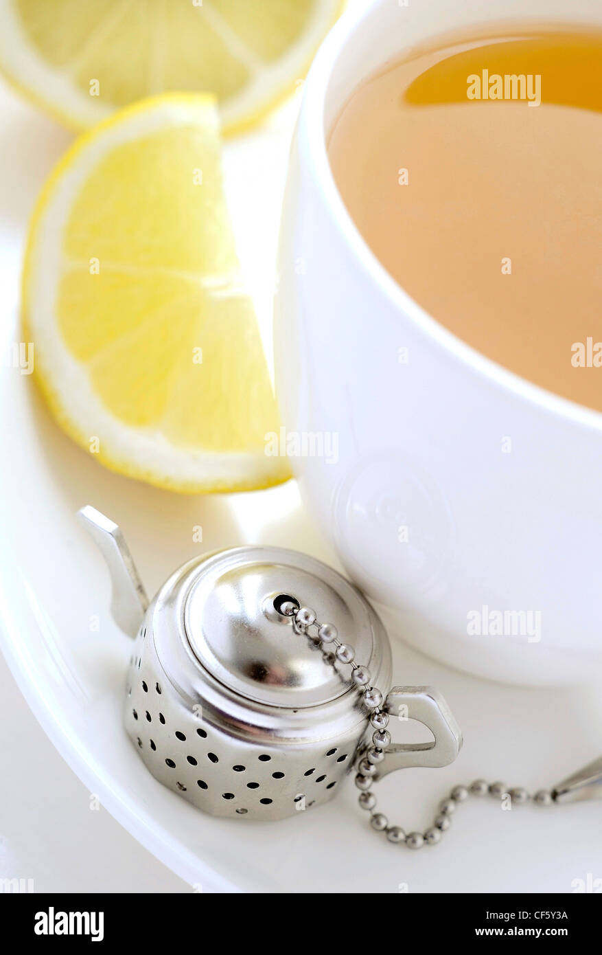 A close up of a white cup and saucer filled a yellow fruit tea, a slice of lemon on one side and a metal teapot shaped infuser Stock Photo