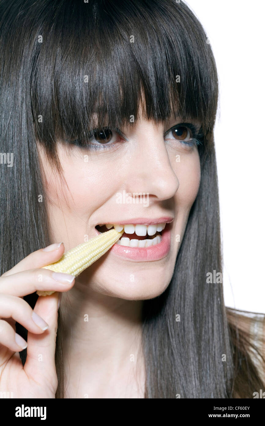 Female long fringed brunette hair, wearing green metallic eyeshadow and pink lipgloss, eating baby corn, smiling, looking up Stock Photo