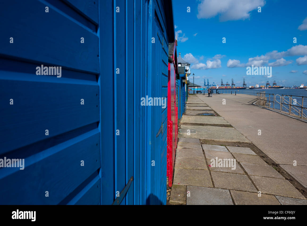 A couple walking arm in arm past recently painted wooden beach huts. Felixstowe Docks are in the background. Stock Photo