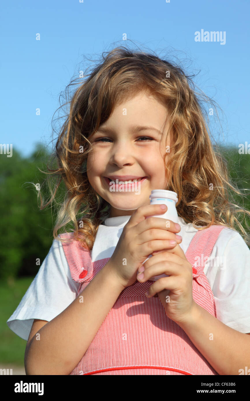 The beautiful little girl with a yoghurt small bottle, against wood and the blue sky. Stock Photo