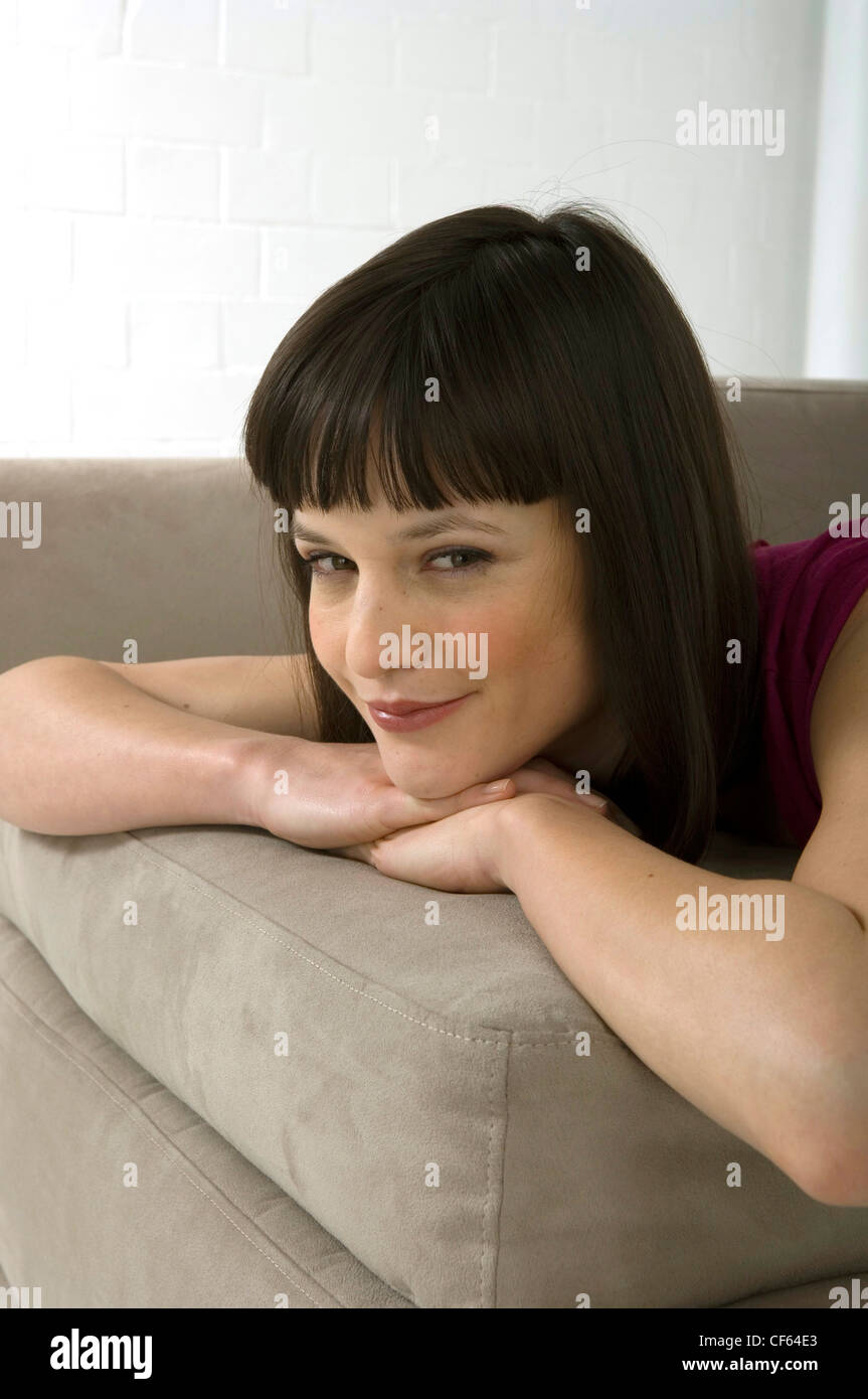 Female with brunette hair in bob lying forward on sofa leaning chin on folded hands looking sideways to camera smiling  RBO Stock Photo