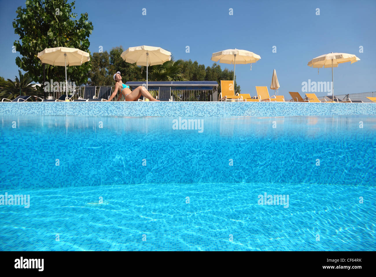Woman becomes tanned near  pool under open-skies in  day-time near deck-chairs and umbrellas, underwater half photo Stock Photo