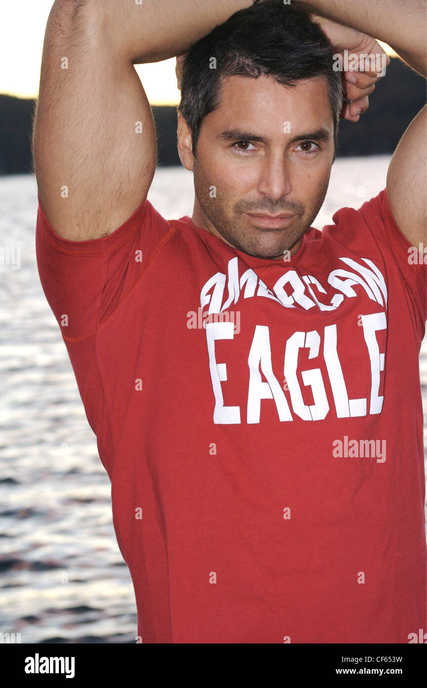 Cropped image of brunette male wearing a red logo T shirt, standing by waters edge arms crossed on top of head Looking to Stock Photo