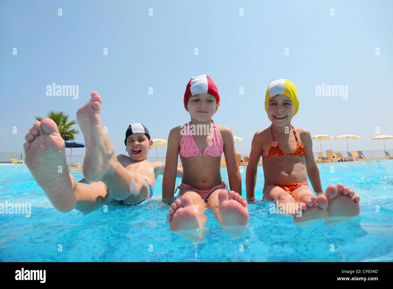 Two girls sit on skirting in  pool and feet overwater and boy lift which falls in water, underwater package shot Stock Photo