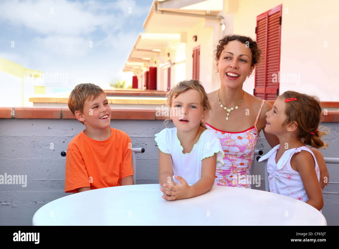 Two girls with boy and mother sit in  day-time on  balcony near  table and speak merrily, focus on little girl in center Stock Photo