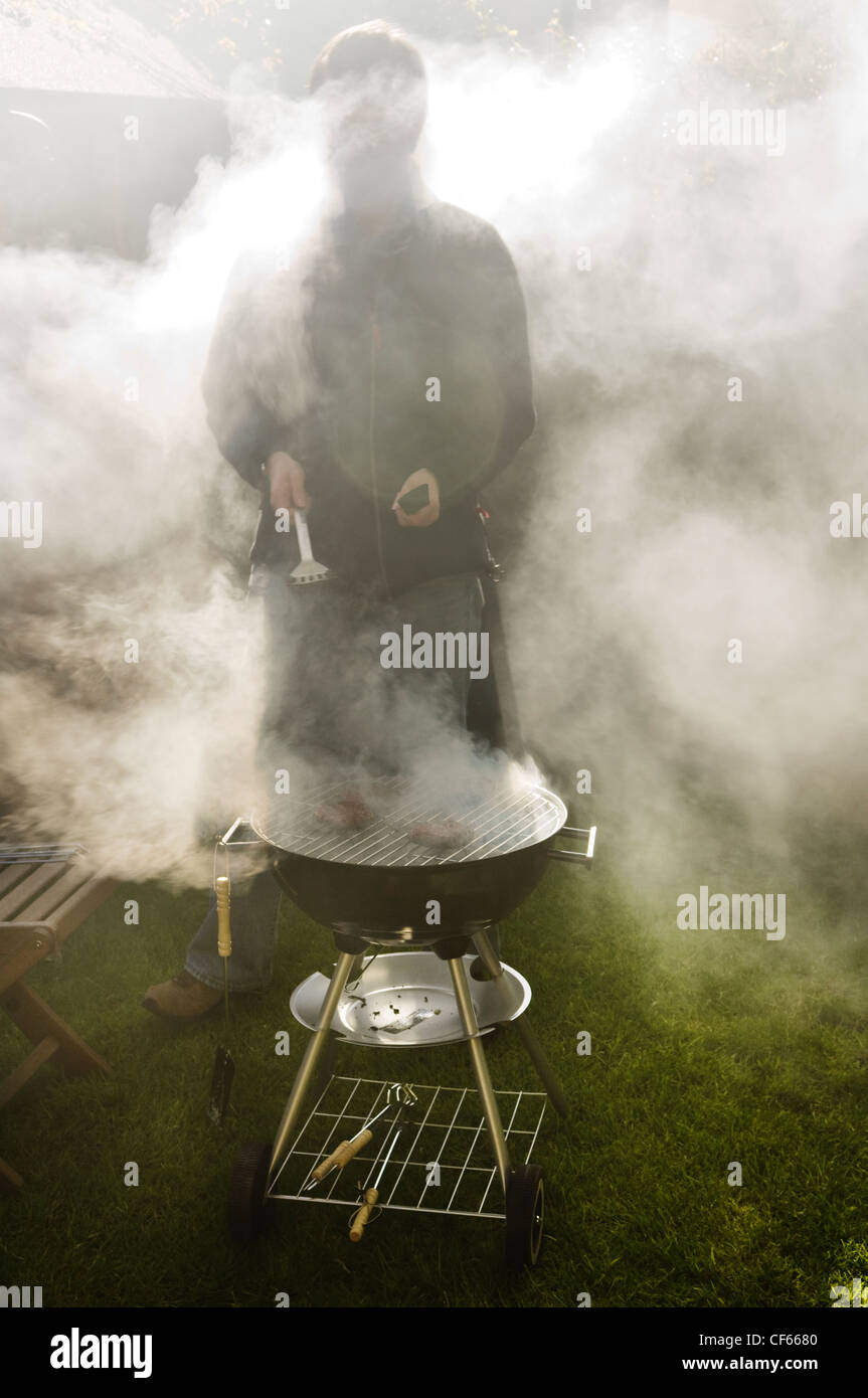 A man shrouded in smoke cooking on a barbecue in a garden. Stock Photo