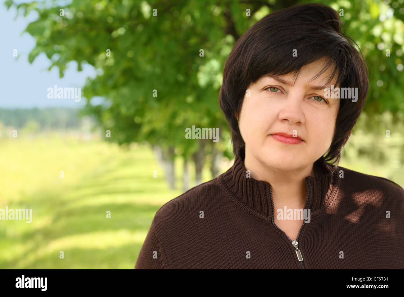 portrait of plumpy adult brunette woman in brown sweater, trees and sky Stock Photo