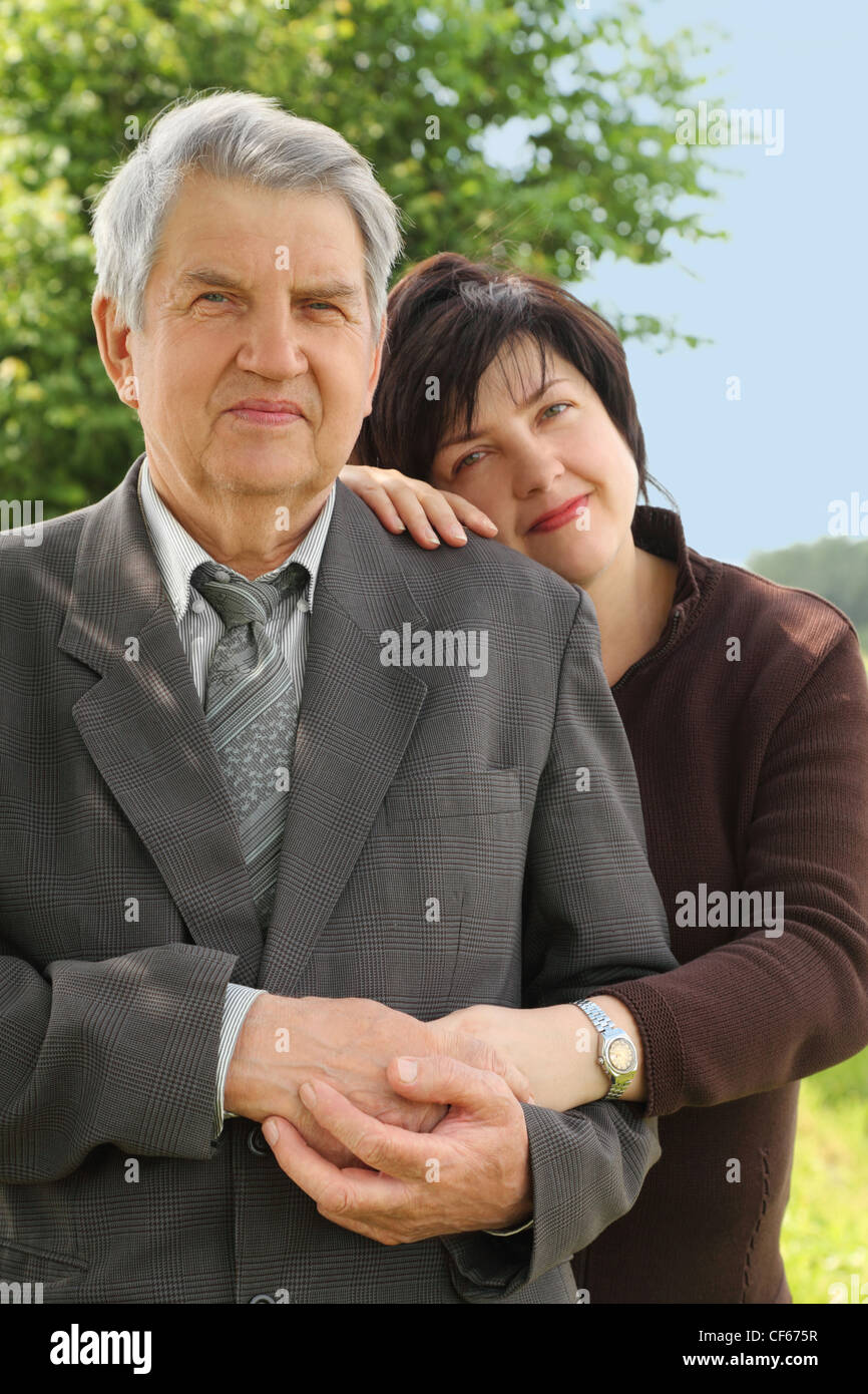 portrait of old senior in suit, his adult daughter leans on his shoulder, smiling, summer trees and sky Stock Photo